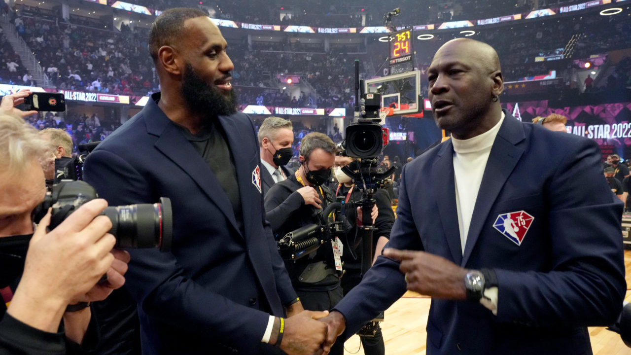 Basketball player LeBron James shaking hands with Michael Jordan on a basketball court surrounded by media members with cameras