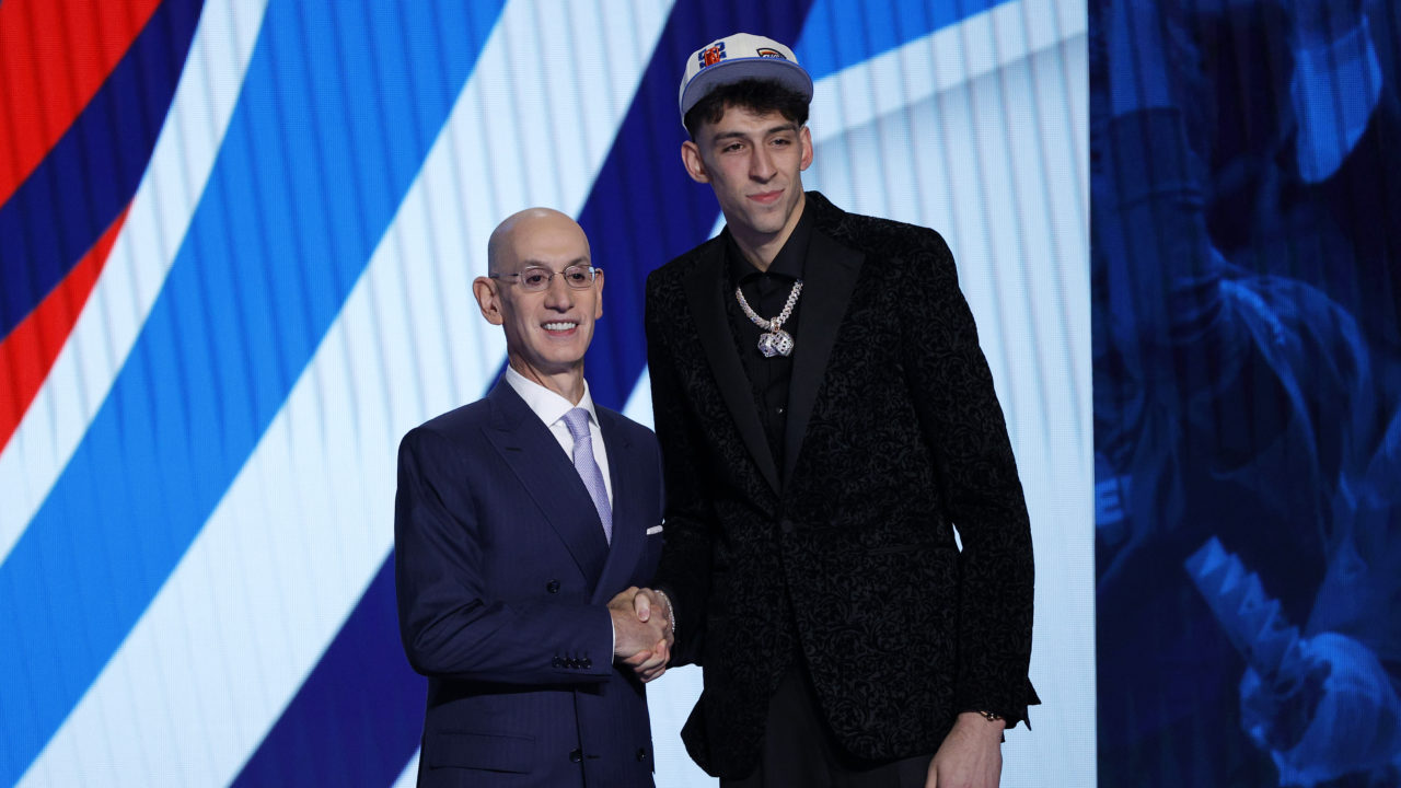 Basketball player Chet Holmgren shaking hands with Adam Silver while standing on a stage and being photographed by cameras