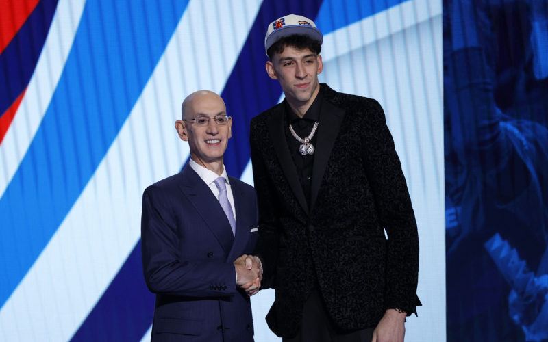 Basketball player Chet Holmgren shaking hands with Adam Silver while standing on a stage and being photographed by cameras