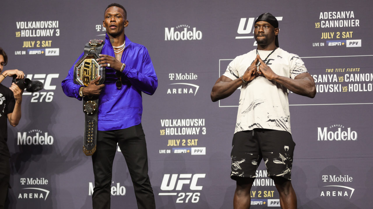 Israel Adesanya standing on a stage with a belt over his shoulder next to Jared Cannonier on his left