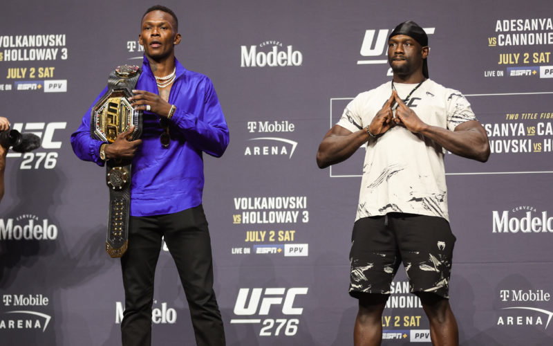 Israel Adesanya standing on a stage with a belt over his shoulder next to Jared Cannonier on his left