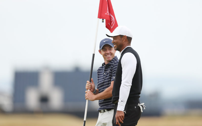 Rory McIlroy holding a golf flag while standing beside Tiger Woods and speaking on a golf course
