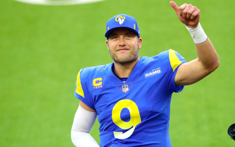 Football player Matthew Stafford smiling and raising his left hand in a thumbs-up on a football field