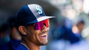 Julio Rodriguez smiles while seated in a baseball dugout while wearing a pair of sunglasses and a Seattle Mariners cap